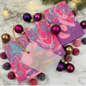 May include: Artisan soap bars with a swirl design in pink, purple, and blue, alongside Christmas ornaments. The soap bars have a tag that reads "Sugar Plum Dreams Artisan Soap". The scene is festive with a marble background and decorative elements.