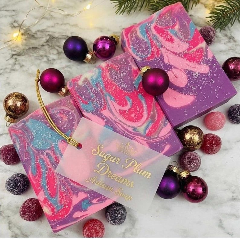 May include: Artisan soap bars with a swirl design in pink, purple, and blue, alongside Christmas ornaments. The soap bars have a tag that reads "Sugar Plum Dreams Artisan Soap". The scene is festive with a marble background and decorative elements.