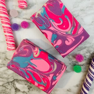 May include: Two rectangular bars of soap with a vibrant swirl pattern in shades of pink, purple, and teal. The soap is surrounded by candy canes and colorful, round candies, creating a festive and colorful display.