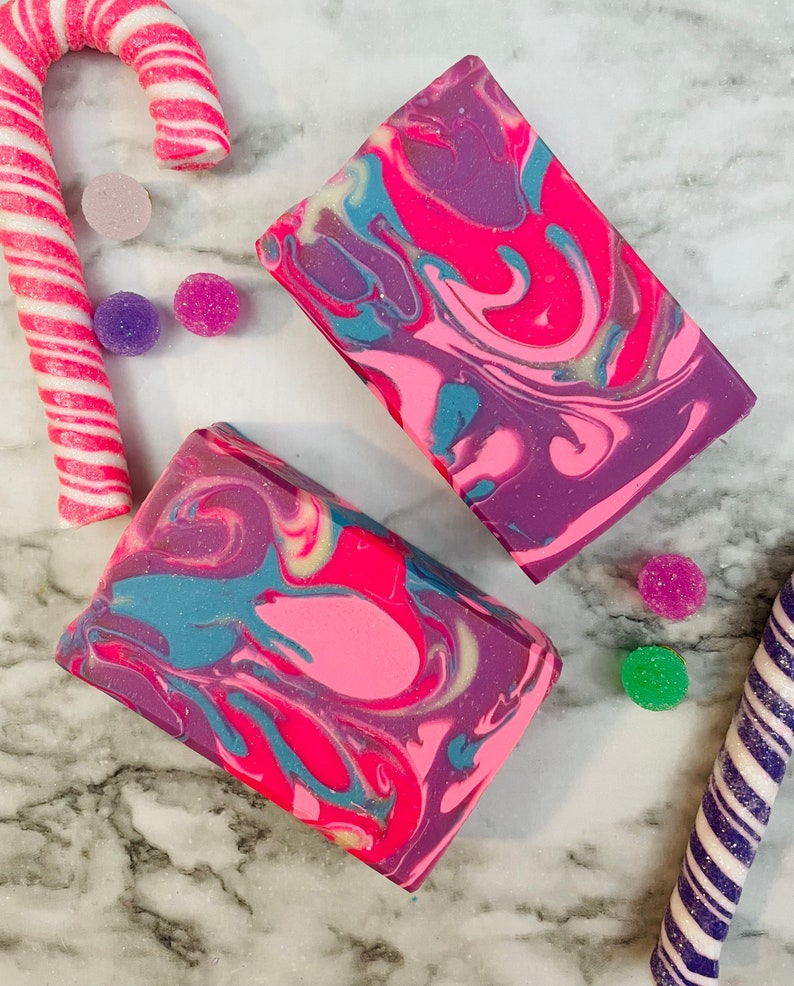 May include: Two rectangular bars of soap with a vibrant swirl pattern in shades of pink, purple, and teal. The soap is surrounded by candy canes and colorful, round candies, creating a festive and colorful display.