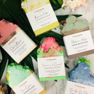 K&ouml;nnte beinhalten: Draufsicht auf sechs handgefertigte Seifen mit verschiedenen Farben und Designs. Jede Seife ist verpackt und mit Namen und Beschreibung versehen. Die Seifen umfassen D&uuml;fte wie Banana Pudding und Save the Sea Turtles. Die Seifen sind auf einer Marmoroberfl&auml;che mit tropischen Bl&auml;ttern angeordnet.