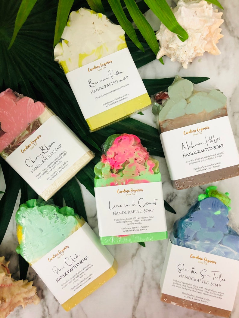 K&ouml;nnte beinhalten: Draufsicht auf sechs handgefertigte Seifen mit verschiedenen Farben und Designs. Jede Seife ist verpackt und mit Namen und Beschreibung versehen. Die Seifen umfassen D&uuml;fte wie Banana Pudding und Save the Sea Turtles. Die Seifen sind auf einer Marmoroberfl&auml;che mit tropischen Bl&auml;ttern angeordnet.