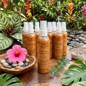 May include: Several amber-coloured Body Glow Shimmer Serum bottles with white spray tops are displayed. A wooden bowl with stones and a pink hibiscus flower is nearby. The bottles have text that reads "Body Glow Shimmer Serum with spf 20".