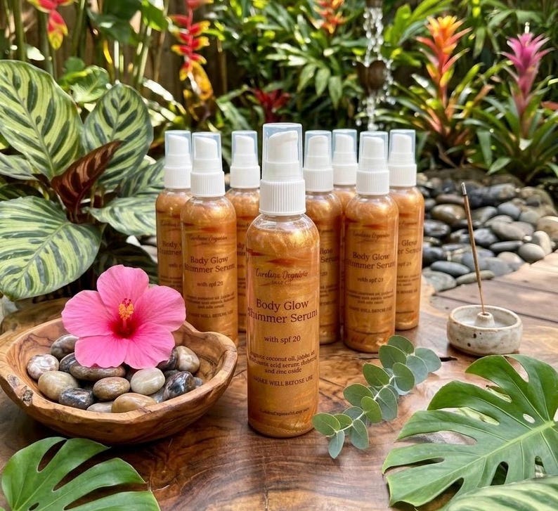 May include: Several amber-coloured Body Glow Shimmer Serum bottles with white spray tops are displayed. A wooden bowl with stones and a pink hibiscus flower is nearby. The bottles have text that reads "Body Glow Shimmer Serum with spf 20".