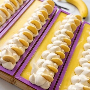 K&ouml;nnte beinhalten: Handgemachte Seifenst&uuml;cke mit gelber Basis und wei&szlig;em, schlagsahne&auml;hnlichem Belag, verziert mit Bananenscheiben. Die Seife befindet sich in lila rechteckigen Formen. Eine frische Banane und Vanilleschoten sind im Hintergrund.