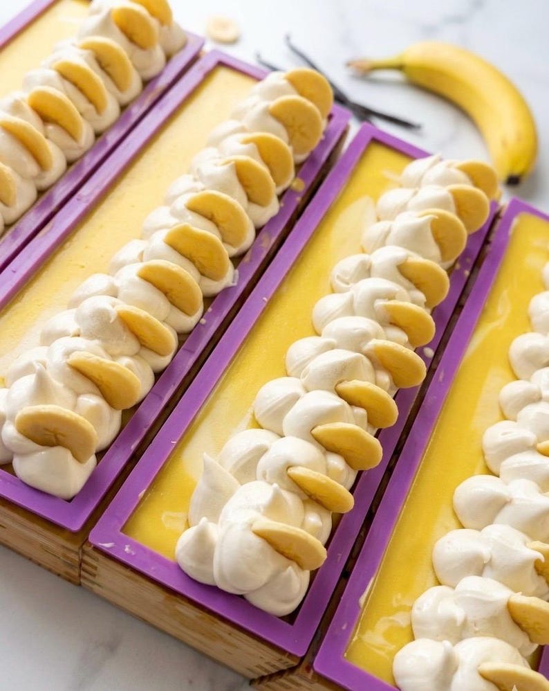 K&ouml;nnte beinhalten: Handgemachte Seifenst&uuml;cke mit gelber Basis und wei&szlig;em, schlagsahne&auml;hnlichem Belag, verziert mit Bananenscheiben. Die Seife befindet sich in lila rechteckigen Formen. Eine frische Banane und Vanilleschoten sind im Hintergrund.