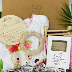 Puede incluir: Un set de regalo para beb&eacute; en una caja blanca. Incluye un letrero de madera con "hello world", un body blanco con dise&ntilde;o de jirafa, un mordedor beige, una pastilla de jab&oacute;n para beb&eacute; y un pa&ntilde;o rosa.