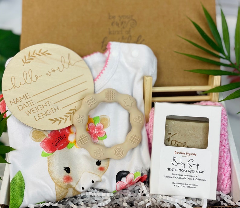 Puede incluir: Un set de regalo para beb&eacute; en una caja blanca. Incluye un letrero de madera con "hello world", un body blanco con dise&ntilde;o de jirafa, un mordedor beige, una pastilla de jab&oacute;n para beb&eacute; y un pa&ntilde;o rosa.