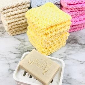 Puede incluir: Una pila de pa&ntilde;os de lavado amarillos de ganchillo junto a una pastilla de jab&oacute;n en un jabonera blanca. El jab&oacute;n est&aacute; estampado con una jirafa y las palabras "Baby Soap". Otras pilas de pa&ntilde;os de lavado crema, azules y rosas est&aacute;n en el fondo.