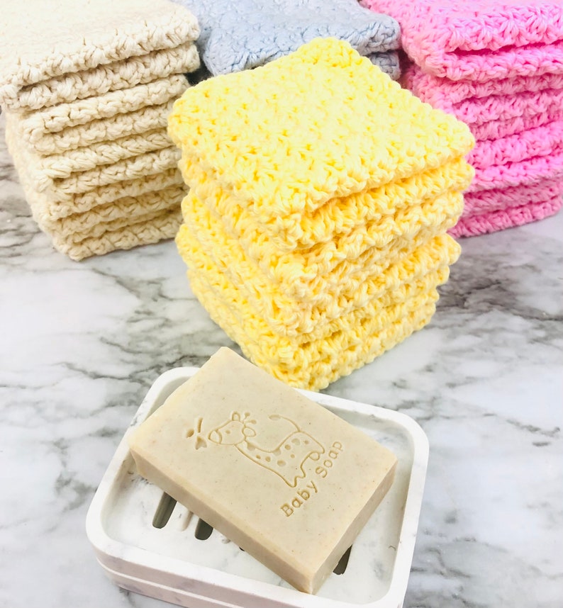 Puede incluir: Una pila de pa&ntilde;os de lavado amarillos de ganchillo junto a una pastilla de jab&oacute;n en un jabonera blanca. El jab&oacute;n est&aacute; estampado con una jirafa y las palabras "Baby Soap". Otras pilas de pa&ntilde;os de lavado crema, azules y rosas est&aacute;n en el fondo.