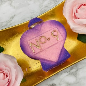 May include: A purple and pink heart-shaped bath bomb with the text "NO. 9" in gold glitter. The bath bomb is on a gold tray, with a pink rose in the background. The bath bomb has a glittery finish, and is likely scented.