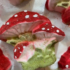 May include: A set of bath bombs shaped like mushrooms. The mushrooms are red and white with green accents. The bath bombs are covered in glitter.