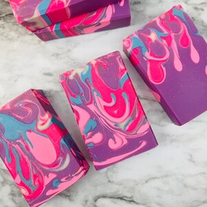 May include: Artisan soap bars with a vibrant swirl design. The soaps feature a blend of colors, including pink, purple, and blue, creating a marbled effect. The bars are rectangular and appear to be handmade, showcasing a unique, colorful pattern.