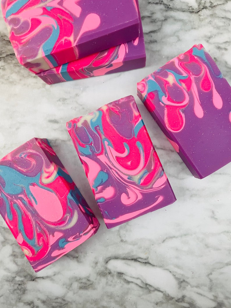 May include: Artisan soap bars with a vibrant swirl design. The soaps feature a blend of colors, including pink, purple, and blue, creating a marbled effect. The bars are rectangular and appear to be handmade, showcasing a unique, colorful pattern.