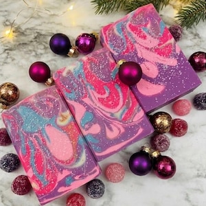 May include: Artisan soap bars with a swirl design in shades of pink, purple, and blue, decorated with glitter. The soaps are surrounded by Christmas ornaments in various colors, including purple, gold, and pink, and a sprig of evergreen.