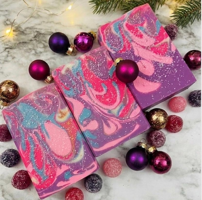 May include: Artisan soap bars with a swirl design in shades of pink, purple, and blue, decorated with glitter. The soaps are surrounded by Christmas ornaments in various colors, including purple, gold, and pink, and a sprig of evergreen.