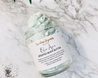 May include: A glass jar of light blue whipped body butter with a white label that reads "Carolina Organics Soap Co. Blue Again Whipped Body Butter. The heart is composed of blue agave flower, sea salt, geranium, and white lily, and the base units orchid, vetiver, cinnamon, musk, and vanilla w/ Shea, Cocoa, & Mango Butters. Cruelty Free & Vegan."