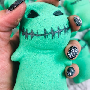 Puede incluir: Una bomba de baño verde con forma de monstruo con una boca cosida y ojos negros.