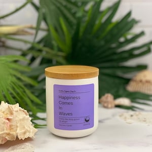 Puede incluir: Vela de cera de soja blanca con tapa de madera. La etiqueta es morada y dice "Happiness Comes In Waves" y "Every little thing gunna be alright".