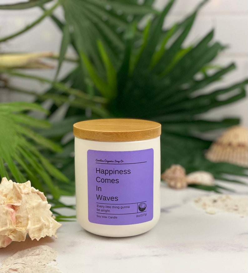 Puede incluir: Vela de cera de soja blanca con tapa de madera. La etiqueta es morada y dice "Happiness Comes In Waves" y "Every little thing gunna be alright".