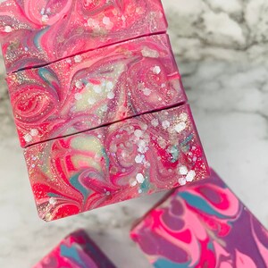 May include: Close-up of handmade soap bars with a swirled design. The soap features vibrant colors of pink, red, and purple, with hints of blue and white. The bars are decorated with glitter, creating a shimmering effect. The soap is a cosmetic product.