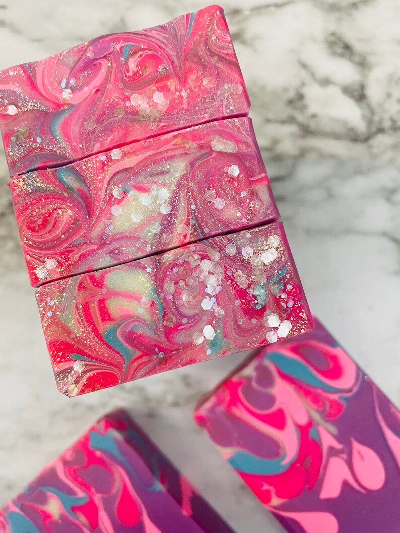 May include: Close-up of handmade soap bars with a swirled design. The soap features vibrant colors of pink, red, and purple, with hints of blue and white. The bars are decorated with glitter, creating a shimmering effect. The soap is a cosmetic product.
