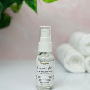 May include: A clear glass bottle with a white pump top containing Hyaluronic Acid Facial Hydration Serum with Aloe + Vitamin B5. The bottle is sitting on a white surface with white towels behind it.