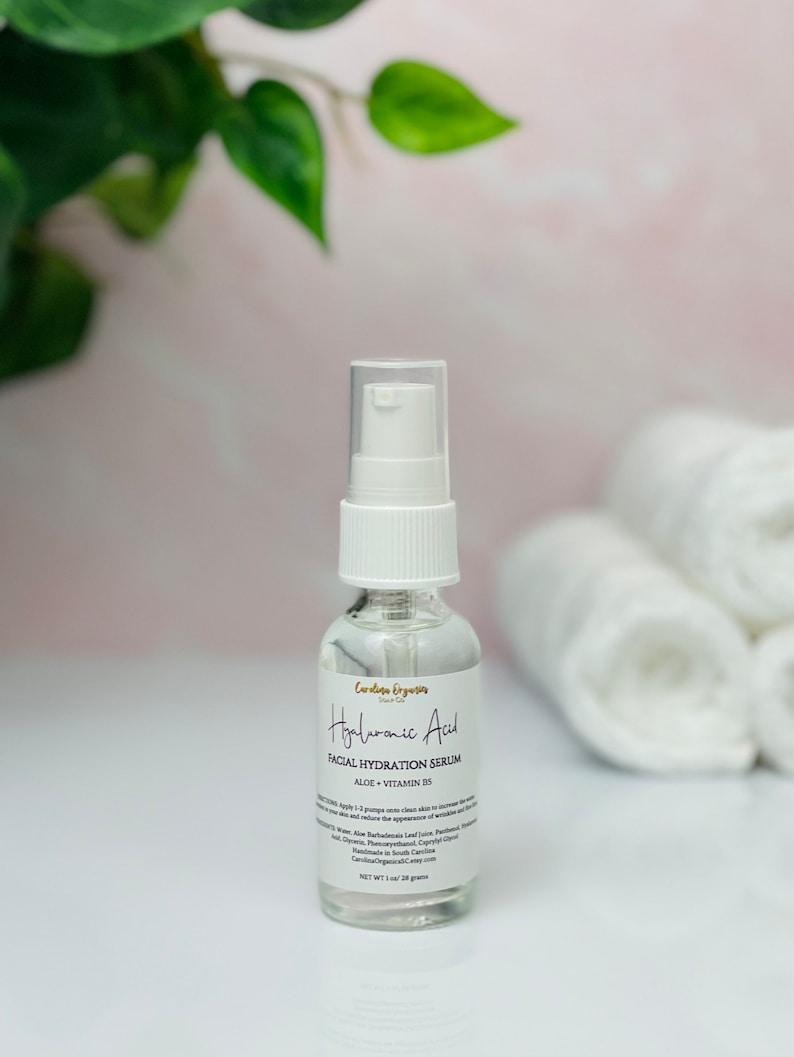 May include: A clear glass bottle with a white pump top containing Hyaluronic Acid Facial Hydration Serum with Aloe + Vitamin B5. The bottle is sitting on a white surface with white towels behind it.