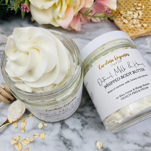 May include: Two glass jars of white whipped body butter with a label that reads "Oatmeal Milk & Honey Whipped Body Butter" with the brand name "Carolina Organics Soap Co." and the text "Notes of oatmeal, milk, honey and almond. w/ Shea, Cocoa, & Mango Butters. Cruelty Free & Vegan."