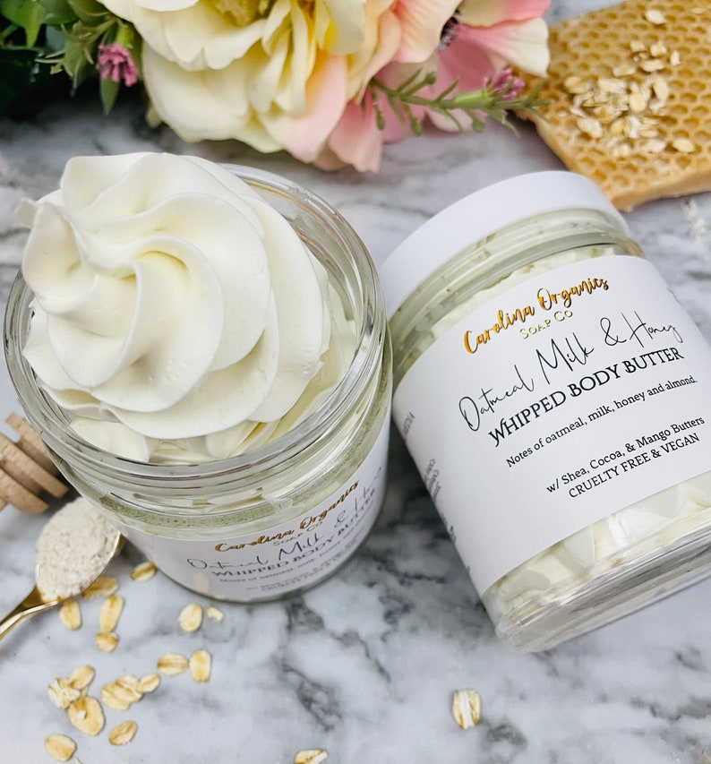 May include: Two glass jars of white whipped body butter with a label that reads "Oatmeal Milk & Honey Whipped Body Butter" with the brand name "Carolina Organics Soap Co." and the text "Notes of oatmeal, milk, honey and almond. w/ Shea, Cocoa, & Mango Butters. Cruelty Free & Vegan."