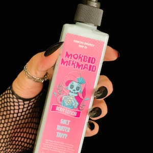 以下が含まれることがあります： 黒いポンプディスペンサー付きのMorbid Mermaidボディローションの長方形ボトル。ピンクのラベルには、骸骨の人魚のイラスト、Salt Water Taffyの文字、成分リスト、正味重量9oz/255グラム、EtsyのURLが記載されています。