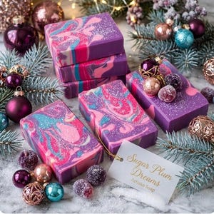 May include: Artisan soap bars with a swirl design in shades of purple, pink, and blue. The bars are decorated with glitter and surrounded by Christmas ornaments and pine branches. A tag reads "Sugar Plum Dreams Artisan Soap."