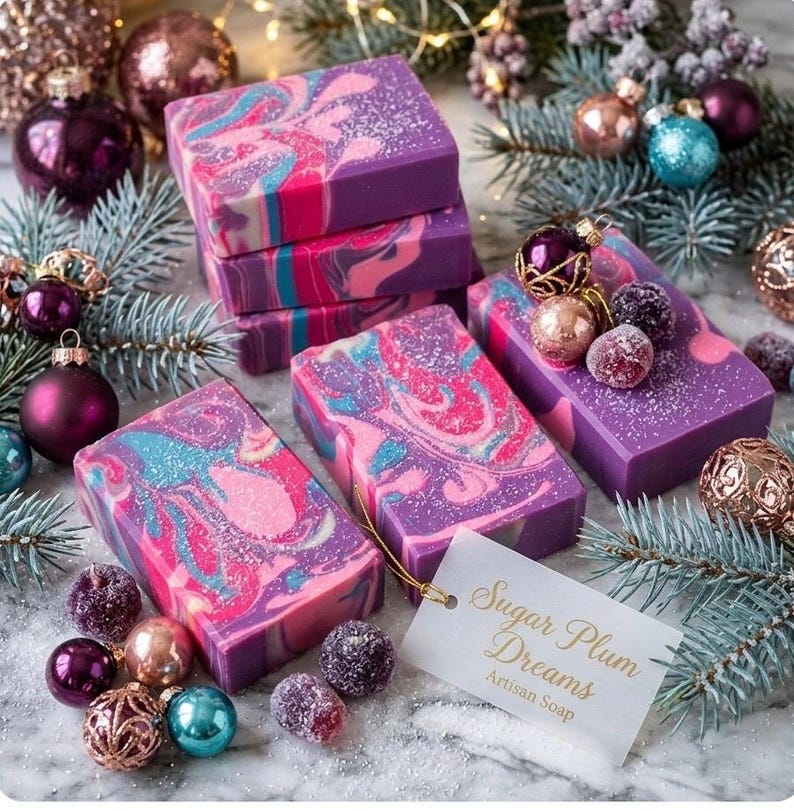 May include: Artisan soap bars with a swirl design in shades of purple, pink, and blue. The bars are decorated with glitter and surrounded by Christmas ornaments and pine branches. A tag reads "Sugar Plum Dreams Artisan Soap."