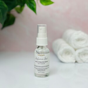 May include: A clear glass bottle with a white pump top containing Hyaluronic Acid Facial Hydration Serum. The bottle is labeled with the product name, ingredients, and net weight of 1 oz (28 grams).