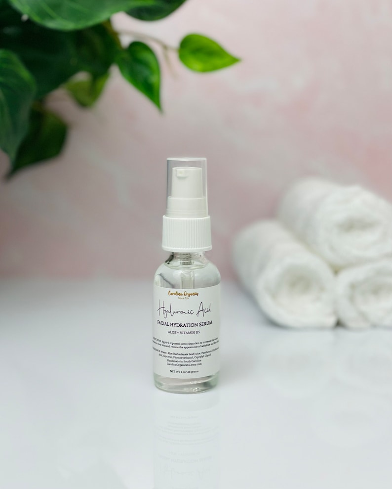 May include: A clear glass bottle with a white pump top containing Hyaluronic Acid Facial Hydration Serum. The bottle is labeled with the product name, ingredients, and net weight of 1 oz (28 grams).