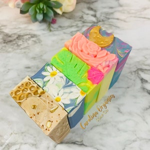 May include: Four handmade soap bars with different designs. The soaps are stacked on top of each other. The top soap is blue with a gold crescent moon design. The second soap is pink with a rose design. The third soap is green with a leaf design. The bottom soap is white with a daisy design. The soaps are labeled "Carolina Organics Soap Co."
