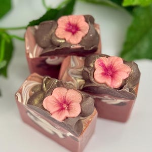 Puede incluir: Cuatro barras rectangulares de jabón artesanal. Cada barra presenta remolinos rosas, marrones y blancos, rematados con una flor rosa y detalles marrones. Los jabones están dispuestos sobre una superficie blanca con hojas verdes de fondo.