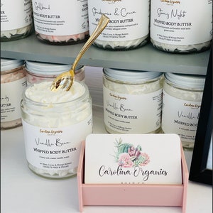 May include: A collection of small glass jars filled with white whipped body butter. The jars have labels with different scents, including "Vanilla Bean", "Strawberry & Cream", "Spallband", and "Stony Night". The jars are arranged on a shelf with a pink business card holder in the front. The business card holder has a floral design and the text "Carolina Organics Soap Co."