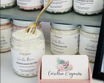 May include: A display of glass jars filled with whipped body butter, labeled with various scents like "Vanilla Bean" and "Strawberries & Cream." A gold spoon rests in one jar. A pink card holder displays cards with a floral design and the text "Carolina Organics."