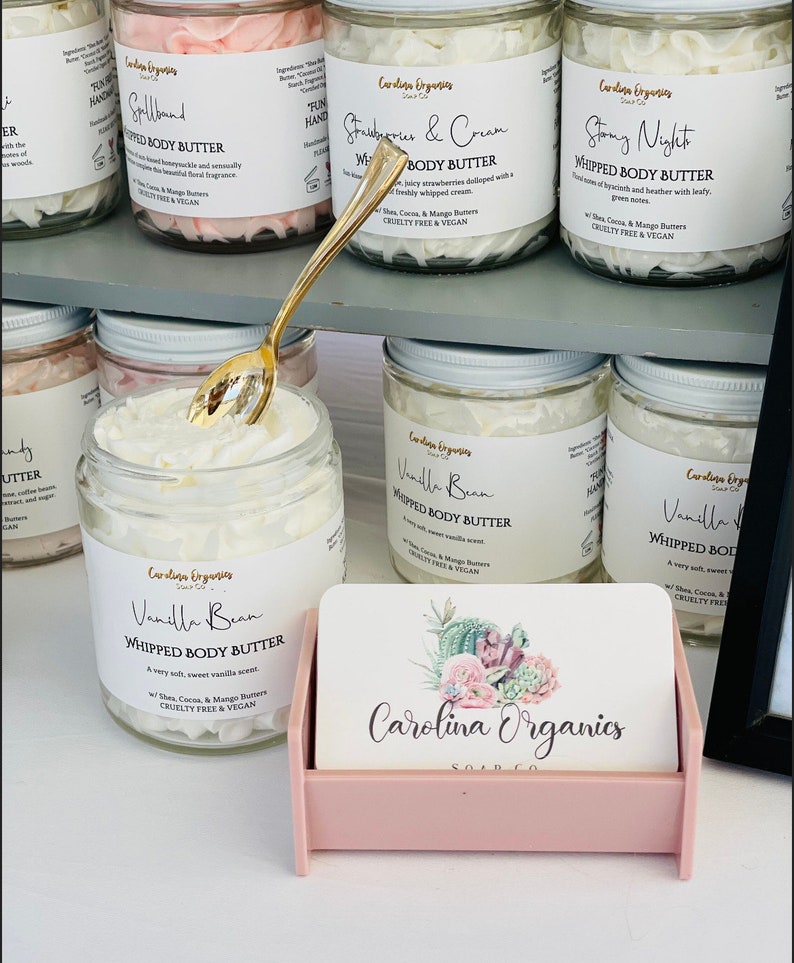 May include: A collection of small glass jars filled with white whipped body butter. The jars have labels with different scents, including "Vanilla Bean", "Strawberry & Cream", "Spallband", and "Stony Night". The jars are arranged on a shelf with a pink business card holder in the front. The business card holder has a floral design and the text "Carolina Organics Soap Co."
