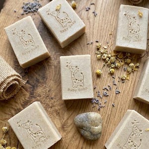 Puede incluir: Varias barras rectangulares de jab&oacute;n para beb&eacute;s, de color marr&oacute;n claro, est&aacute;n dispuestas sobre una superficie de madera. Cada barra est&aacute; estampada con un dise&ntilde;o de jirafa y la inscripci&oacute;n "Baby Soap". Flores secas y lavanda dispersas.