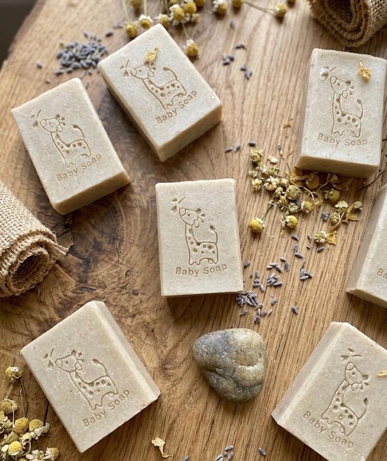 Puede incluir: Varias barras rectangulares de jab&oacute;n para beb&eacute;s, de color marr&oacute;n claro, est&aacute;n dispuestas sobre una superficie de madera. Cada barra est&aacute; estampada con un dise&ntilde;o de jirafa y la inscripci&oacute;n "Baby Soap". Flores secas y lavanda dispersas.