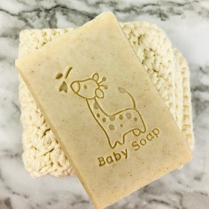 Puede incluir: Una pastilla rectangular de jab&oacute;n para beb&eacute;s con un dise&ntilde;o de jirafa y las palabras "Baby Soap" grabadas en la superficie. El jab&oacute;n descansa sobre un pa&ntilde;o de lavado de ganchillo de color crema. El fondo es una superficie de m&aacute;rmol.