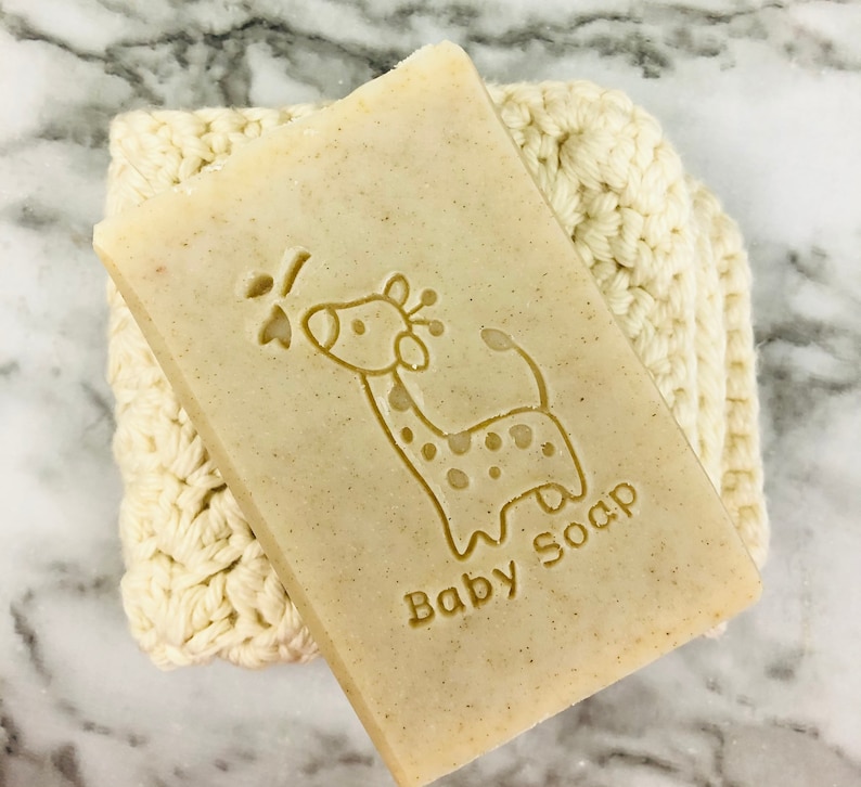 Puede incluir: Una pastilla rectangular de jab&oacute;n para beb&eacute;s con un dise&ntilde;o de jirafa y las palabras "Baby Soap" grabadas en la superficie. El jab&oacute;n descansa sobre un pa&ntilde;o de lavado de ganchillo de color crema. El fondo es una superficie de m&aacute;rmol.