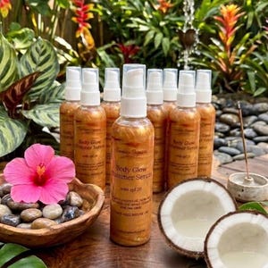 May include: Several bottles of Body Glow Shimmer Serum with SPF 20, alongside a pink hibiscus flower, a wooden bowl of pebbles, and halved coconuts. The bottles have a golden shimmer and white spray tops.