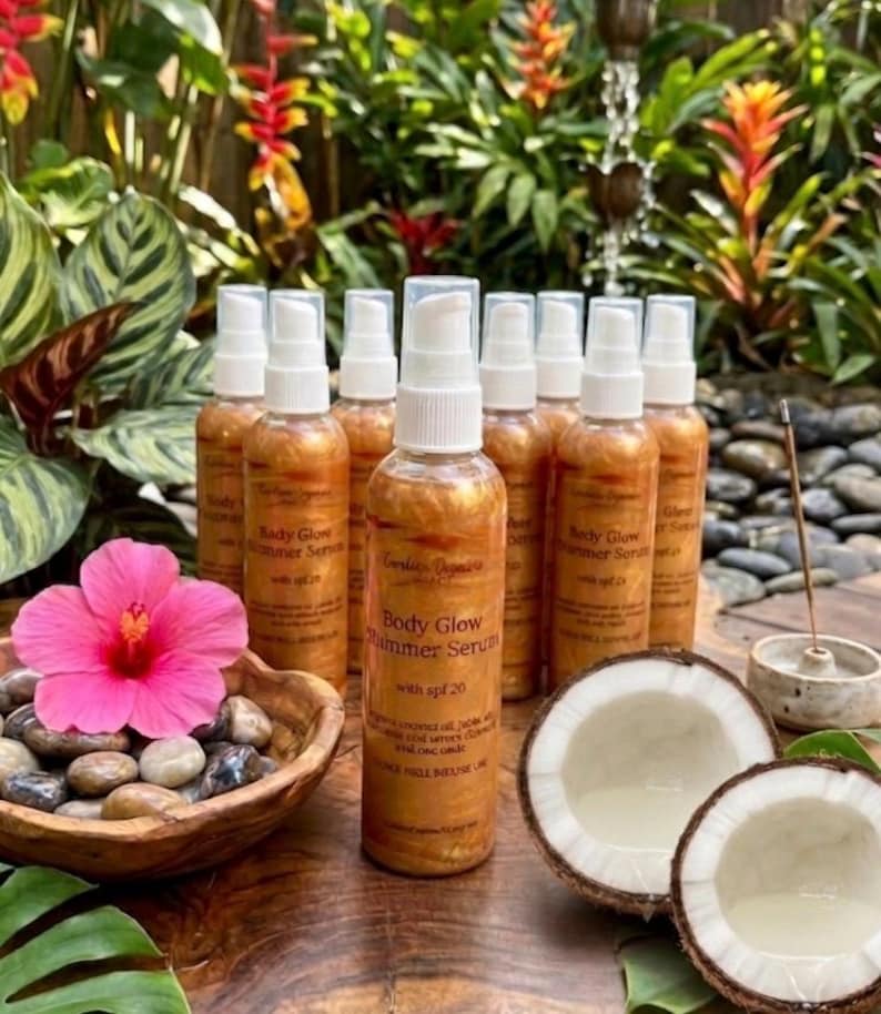 May include: Several bottles of Body Glow Shimmer Serum with SPF 20, alongside a pink hibiscus flower, a wooden bowl of pebbles, and halved coconuts. The bottles have a golden shimmer and white spray tops.
