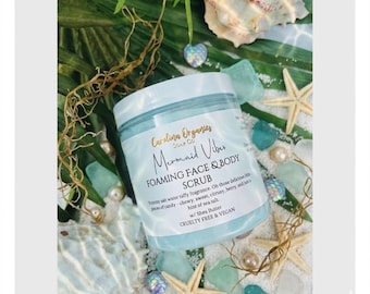 May include: A jar of "Mermaid Vibes" foaming face and body scrub by Carolina Organics. The jar is light blue with a white lid, surrounded by starfish, sea glass, and shells. The label describes the scent as taffy, candy, citrus, berry, and sea salt.