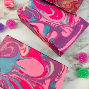 May include: Close-up of three rectangular bars of handmade soap with a swirled design in shades of pink, purple, and blue. The soap is surrounded by colorful candies and sugar-coated spheres, suggesting a festive theme.