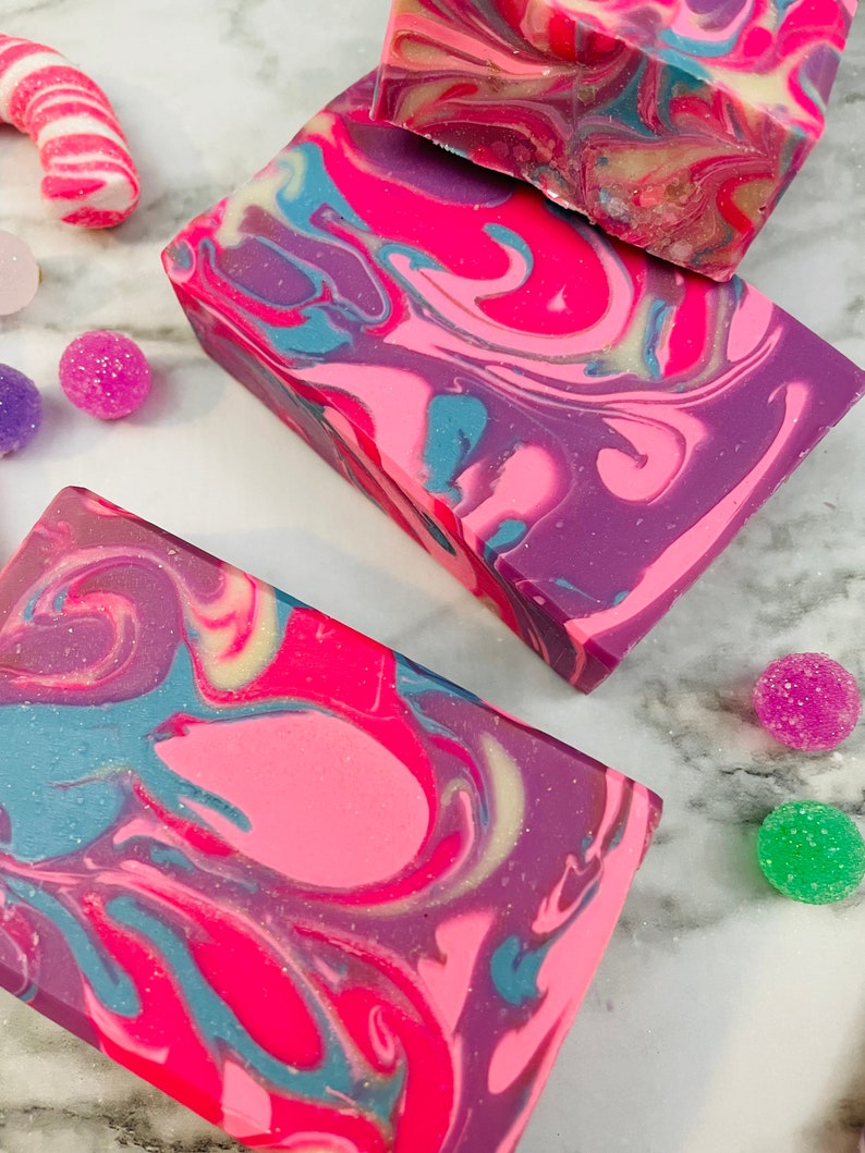 May include: Close-up of three rectangular bars of handmade soap with a swirled design in shades of pink, purple, and blue. The soap is surrounded by colorful candies and sugar-coated spheres, suggesting a festive theme.