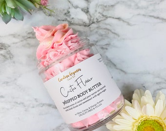 May include: A clear glass jar filled with pink whipped body butter, labeled "Cactus Flower." The label details the product's ingredients and features. The jar is surrounded by flowers on a marble surface.